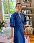 A woman standing in a room with bookshelves wearing a royal blue dress with puff sleeves, a V-neck, and a sash belt.