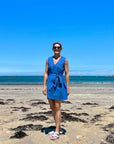 A woman standing on a beach wearing a sleeveless, trapeze-shaped royal blue mini dress with a deep v-neck, paired with a sash belt.