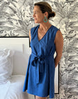 A woman standing in a bedroom wearing a sleeveless, trapeze-shaped royal blue mini dress with a deep v-neck, paired with a sash belt.