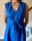 A woman wearing a sleeveless, trapeze-shaped royal blue mini dress with a deep v-neck, paired with a sash belt.