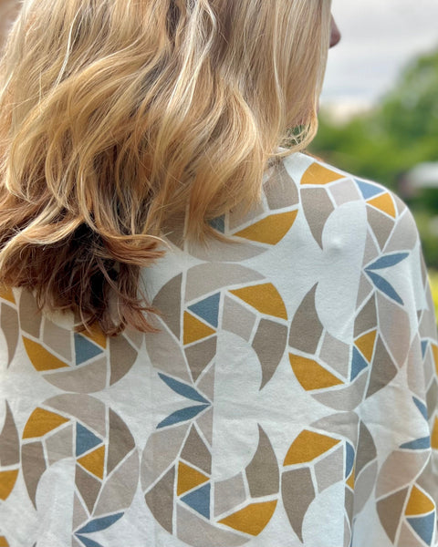 A person wearing a cream-colored cozy wrap/scarf with a geometric pattern featuring mustard yellow, taupe, and gray blue