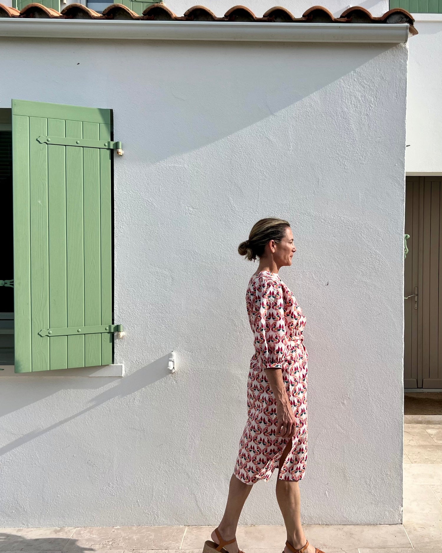 Woman in a patterned dress walking past a white building with green shutters.