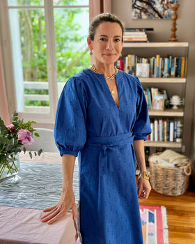 A woman standing in a room with bookshelves wearing a royal blue dress with puff sleeves, a V-neck, and a sash belt.