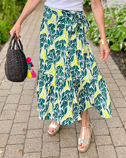A person wearing a chartreuse snake ruffle wrap skirt with a dark teal green and chartreuse print holding a black straw bag standing on a pathway 