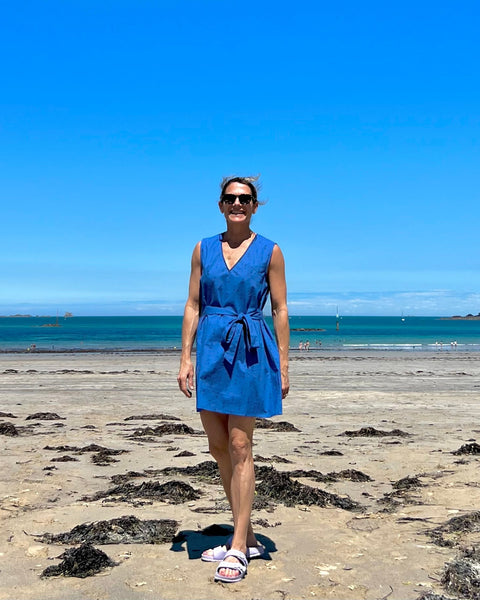 A woman standing on a beach wearing a sleeveless, trapeze-shaped royal blue mini dress with a deep v-neck, paired with a sash belt. 