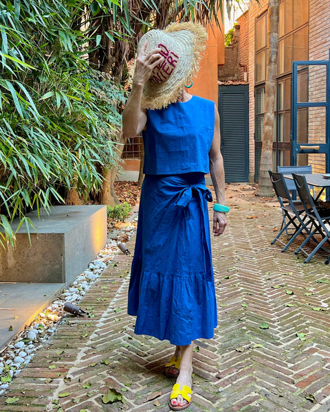 A person wearing a blue sleeveless crew neck crop top with a blue wrap skirt with a ruffle on a patio in Italy
