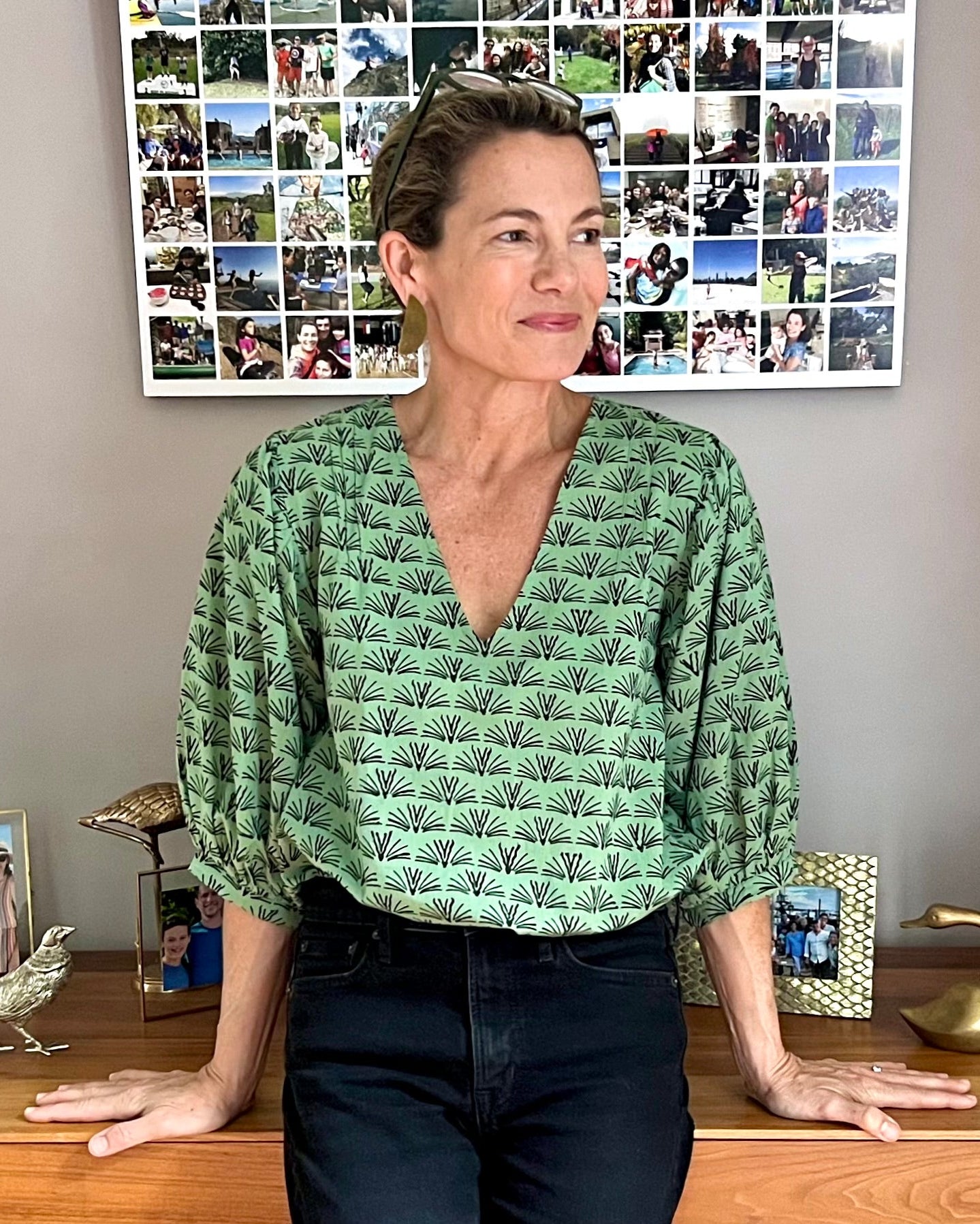 A woman wearing a pine green colored blouse with a blackbuck animal pattern and puffy sleeves, standing in a room with photo frames and a lamp in the background.