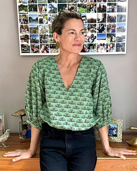 A woman wearing a pine green colored blouse with a blackbuck animal pattern and puffy sleeves, standing in a room with photo frames and a lamp in the background.