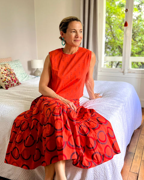 A person seated on a bed wearing a sleeveless, red and purple print, boxy top with a crew neck.