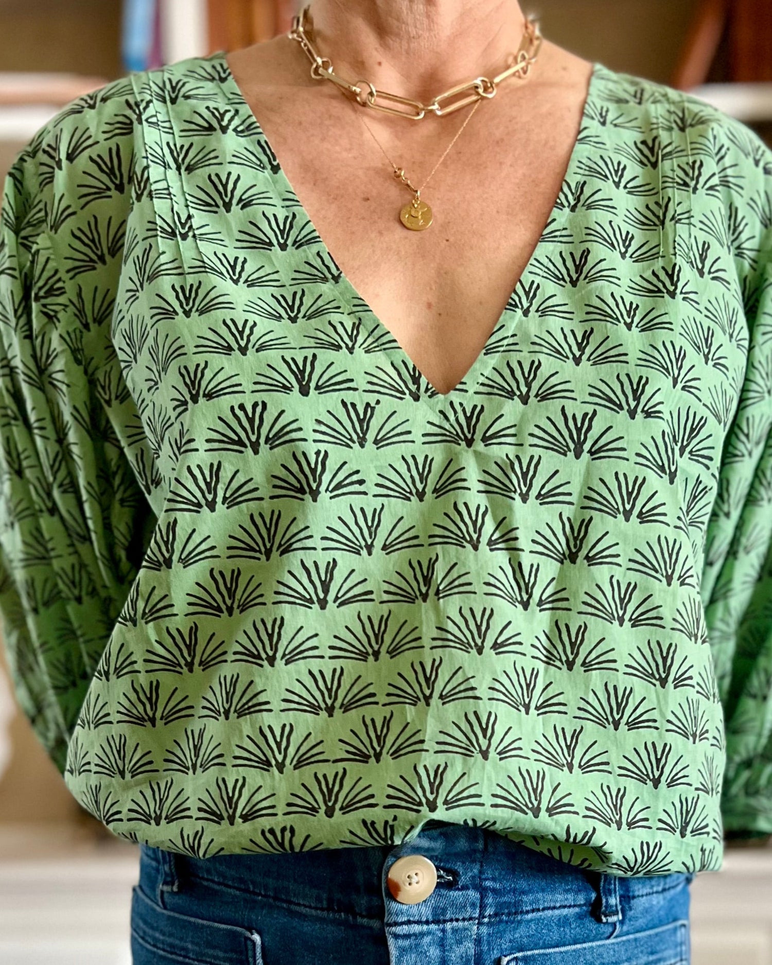 A woman wearing a pine green colored blouse with a blackbuck animal pattern and puffy sleeves, standing in a room with bookshelves.