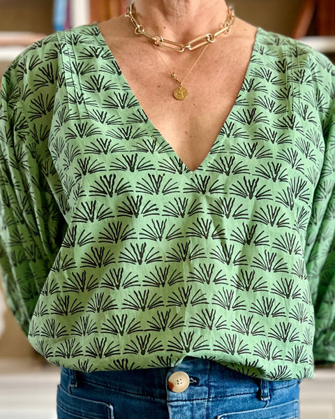 A woman wearing a pine green colored blouse with a blackbuck animal pattern and puffy sleeves, standing in a room with bookshelves.