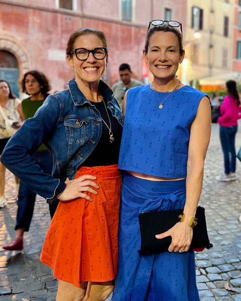 A person wearing a blue sleeveless crew neck crop and blue wrap skirt with her friend wearing a red wrap skirt and denim jacket in piazza in Rome