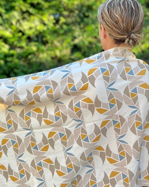 A person wearing a cream-colored cozy wrap/scarf with a geometric pattern featuring mustard yellow, taupe, and gray blue, standing in front of a bush 