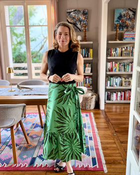 A woman standing in a beautifully decorated living room wearing a green wrap skirt with a ruffle, paired with a black top and sandals.