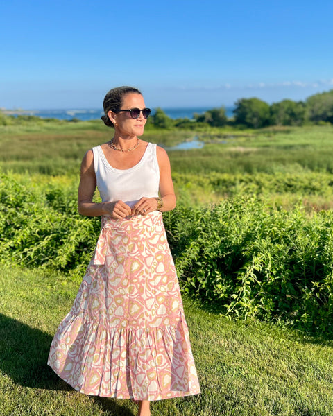 A woman standing on a grassy field with a blush pink and khaki animal print wrap skirt with a ruffle, paired with a white top and sunglasses.