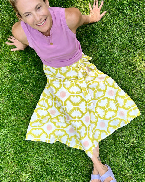 A person seated on grass wearing a wrap skirt with a geometric pattern in yellow and pink on an off-white background