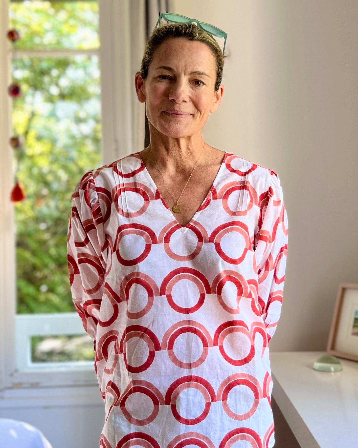 A person wearing a white blouse with a coral and pink geometric pattern, featuring a deep-V neck and puff sleeves in front of a window in a sun-filled bedroom.