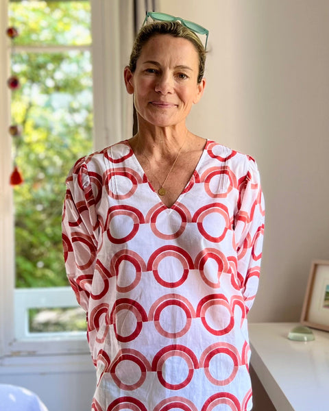 A person wearing a white blouse with a coral and pink geometric pattern, featuring a deep-V neck and puff sleeves in front of a window in a sun-filled bedroom.