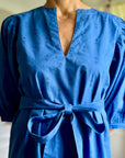 A woman standing in a room wearing a royal blue dress with puff sleeves, a V-neck, and a sash belt.