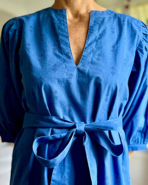 A woman standing in a room wearing a royal blue dress with puff sleeves, a V-neck, and a sash belt.