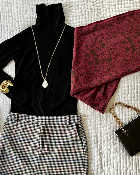A flat lay outfit with a wine colored coazy wrap, wool skirt, black turtleneck, gold jewelry, and a black purse