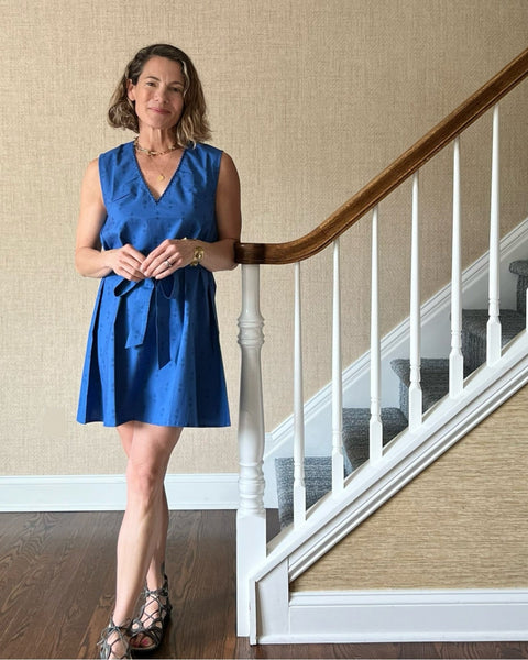 A woman standing by stairs wearing a sleeveless, trapeze-shaped royal blue mini dress with a deep v-neck, paired with a sash belt. 