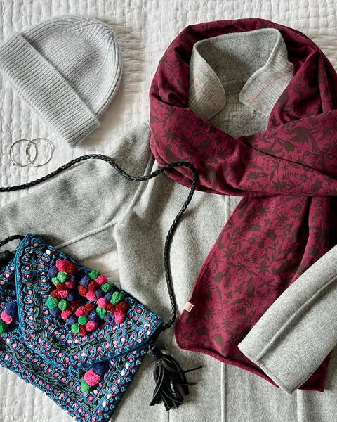 A flat lay outfit with a wine colored wrap/scarf, gray wool hat, gray wool coat, and multi-colored purse 