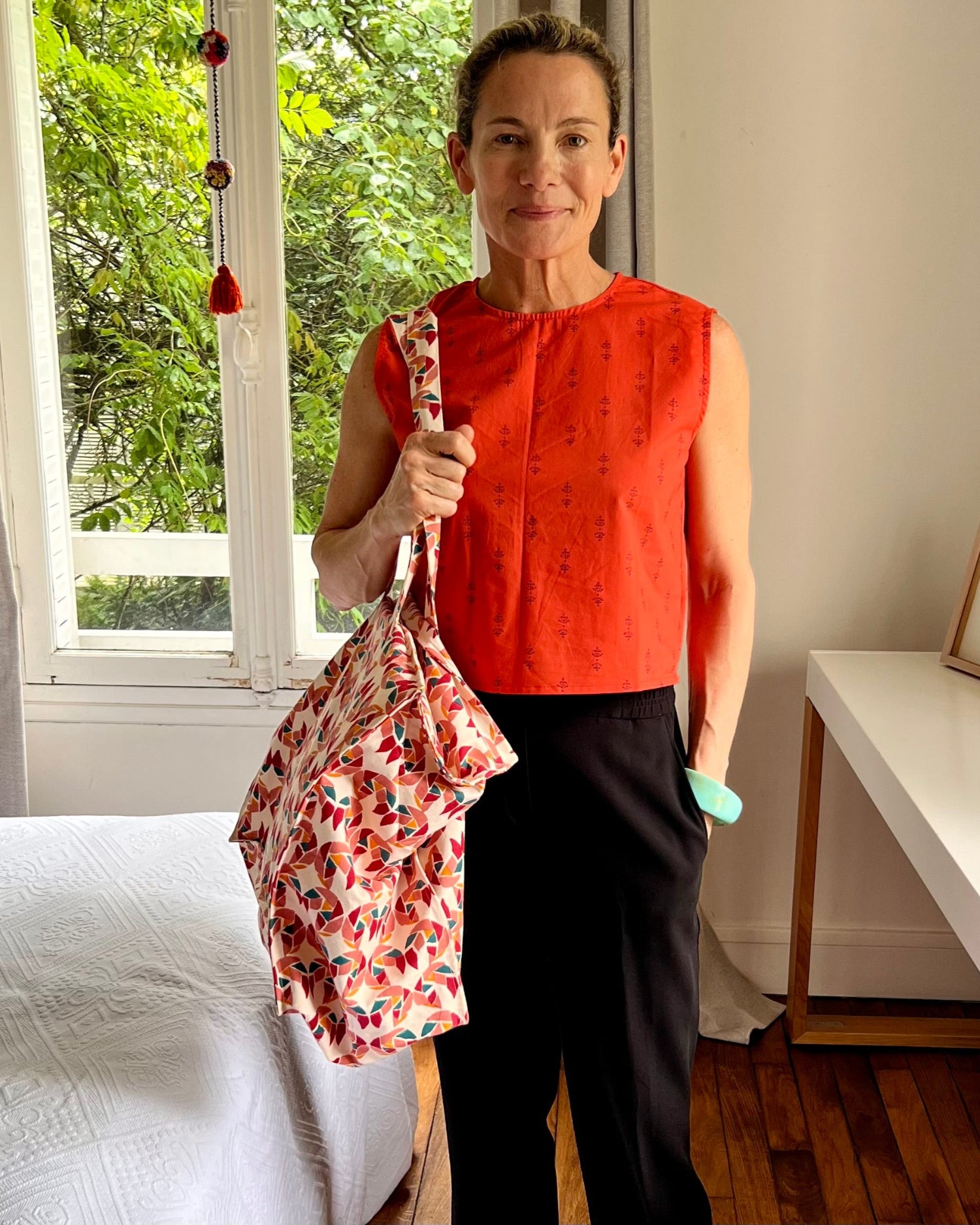 A person wearing a sleeveless, red and purple print, boxy top with a crew neck and black pants holding a brightly colored tote.
