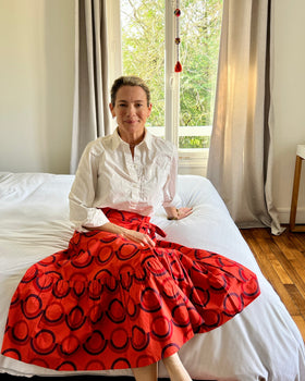 A person sitting on a bed wearing a red and purple geometric patterned skirt with a wrap-around style.