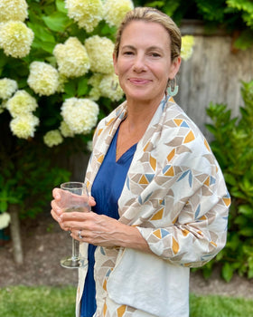 A person wearing a cream-colored cozy wrap/scarf with a geometric pattern featuring mustard yellow, taupe, and gray blue, standing in front of a bush with white flowers.