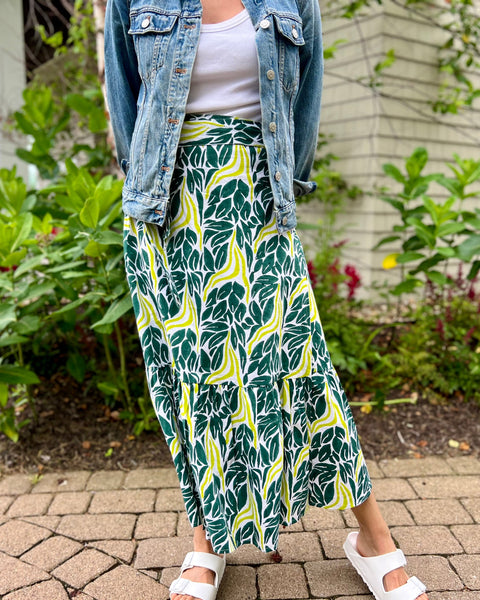 A person wearing a chartreuse snake ruffle wrap skirt with a dark teal green and chartreuse print, paired with a denim jacket, standing on a pathway with grass and buildings in the background.