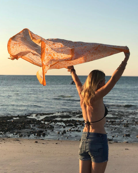 Woman on a beach holding an orange hand block-printed jaguar sarong / grand scarf over her head in the wind with ocean and sunset in the background