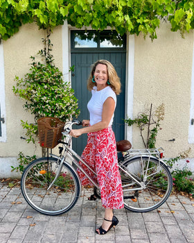 Woman wearing beautiful red and purple block-printed wrap skirt standing next to a bicycle in front of a building with greenery