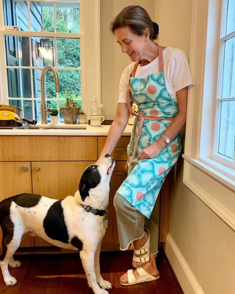Woman wearing a turquoise and terracotta hand block-printed apron, standing in a kitchen with a dog