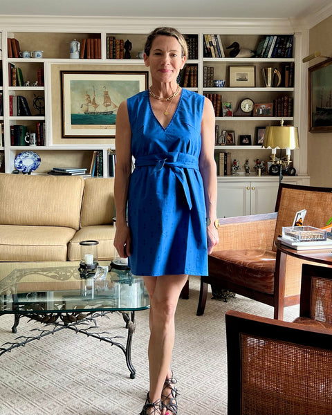A woman standing in a living room wearing a sleeveless, trapeze-shaped royal blue mini dress with a deep v-neck, paired with a sash belt. 