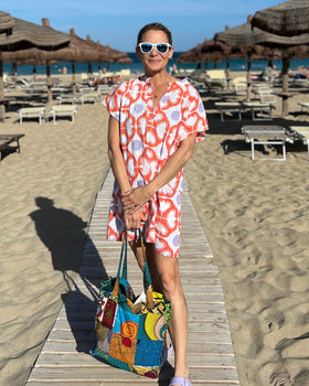 Woman on a beach wearing sunglasses and a coral, lavender, and white hand block-printed short kaftan dress holding a bag