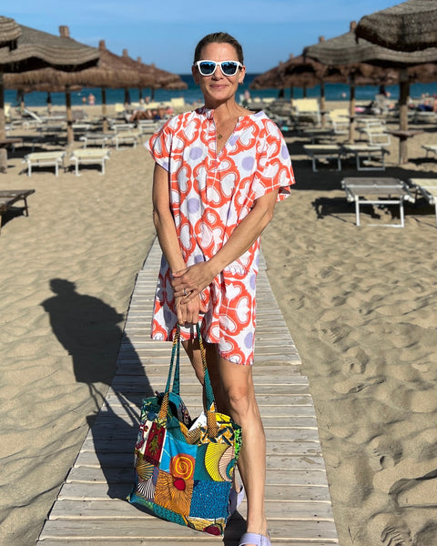 Woman on a beach wearing sunglasses and a coral, lavender, and white hand block-printed short kaftan dress holding a bag