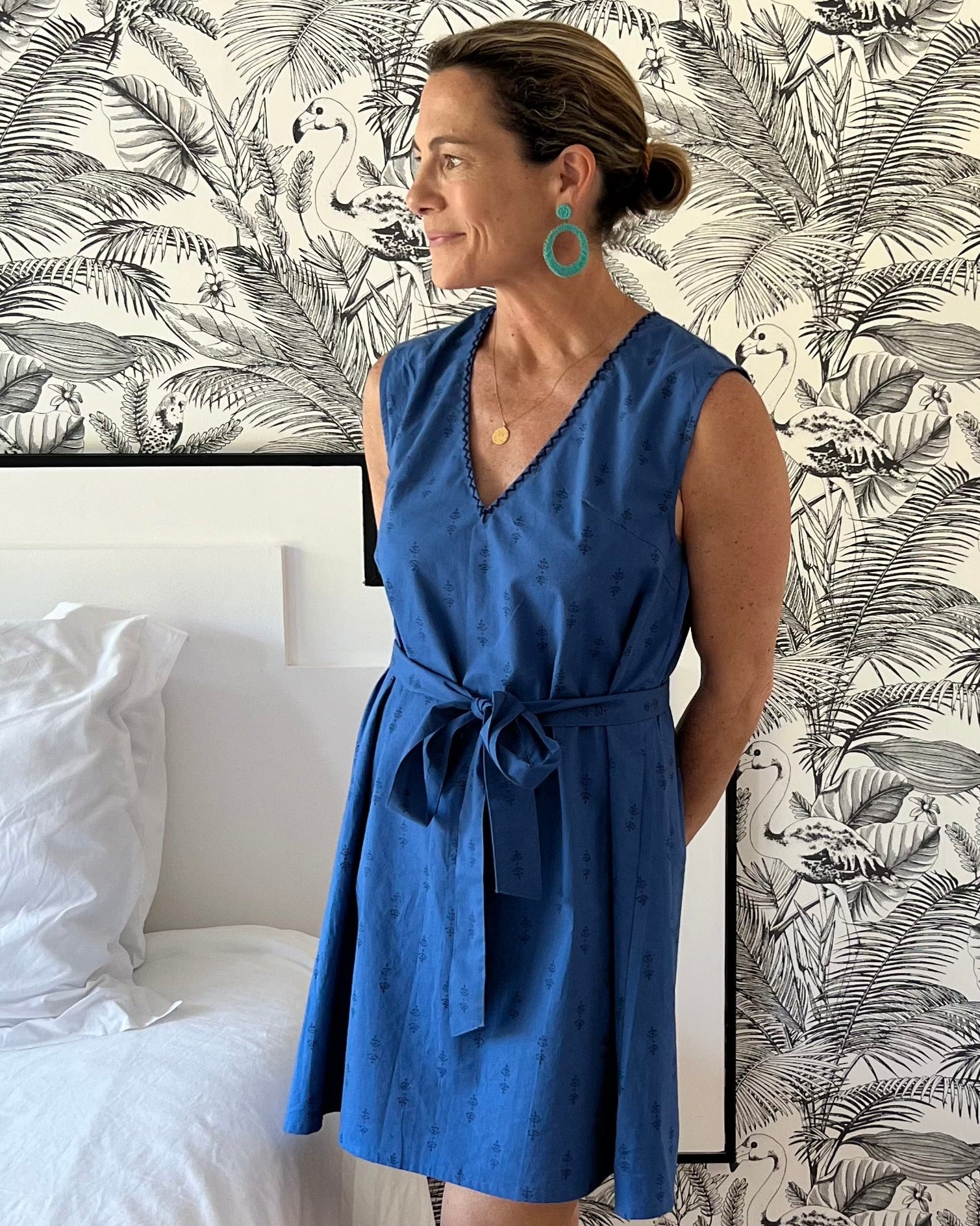 A woman standing in a bedroom wearing a sleeveless, trapeze-shaped royal blue mini dress with a deep v-neck, paired with a sash belt. 