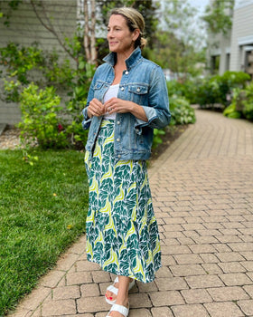 A person wearing a chartreuse snake ruffle wrap skirt with a dark teal green and chartreuse print, paired with a denim jacket, standing on a pathway with grass and buildings in the background.
