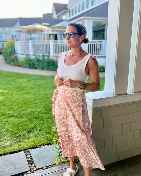 A woman standing on a porch with a blush pink and khaki animal print wrap skirt with a ruffle, paired with a white top and sunglasses.