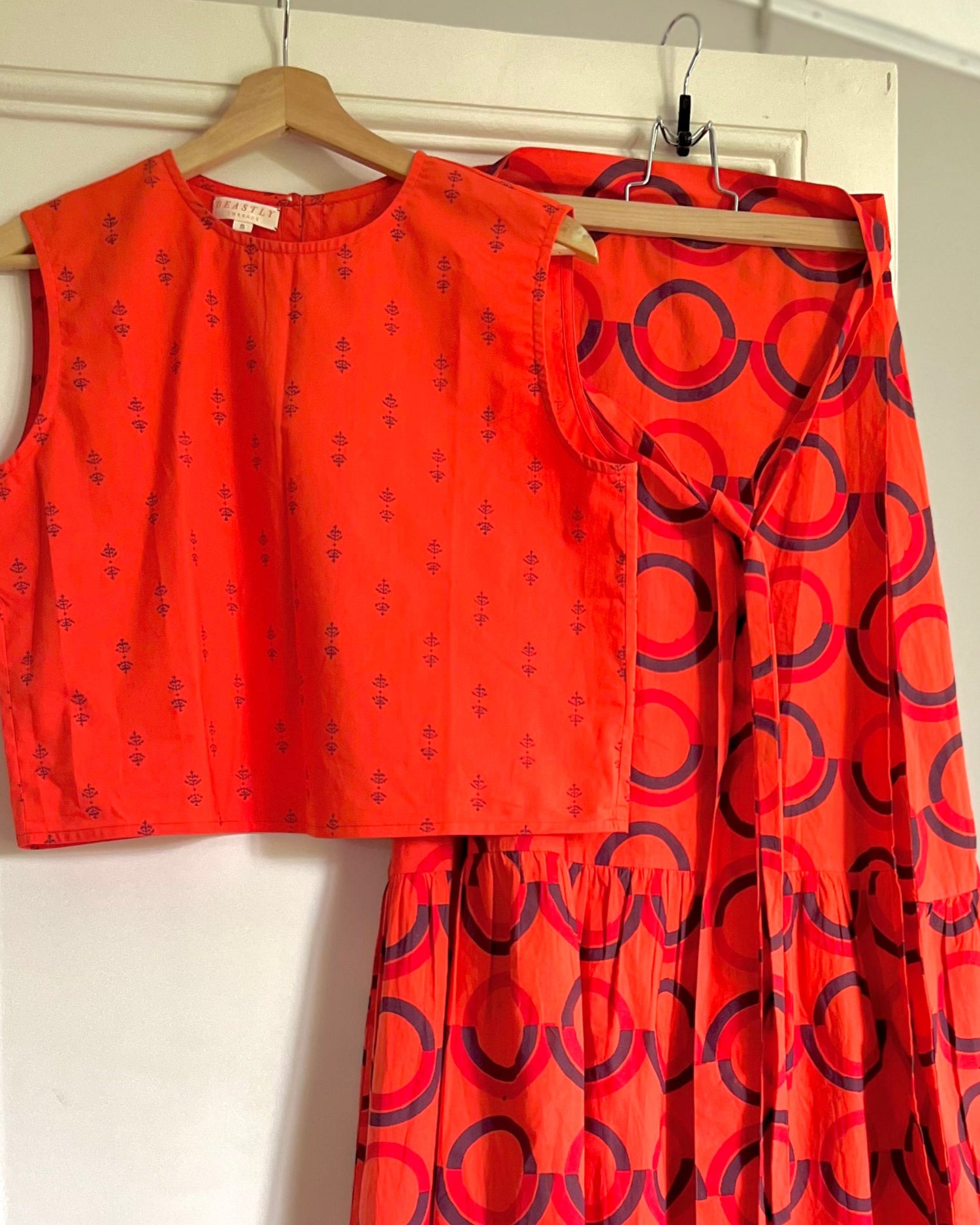 A sleeveless, red and purple print, boxy top with a crew neck and a red block printed wrap skirt with a ruffle on hangers