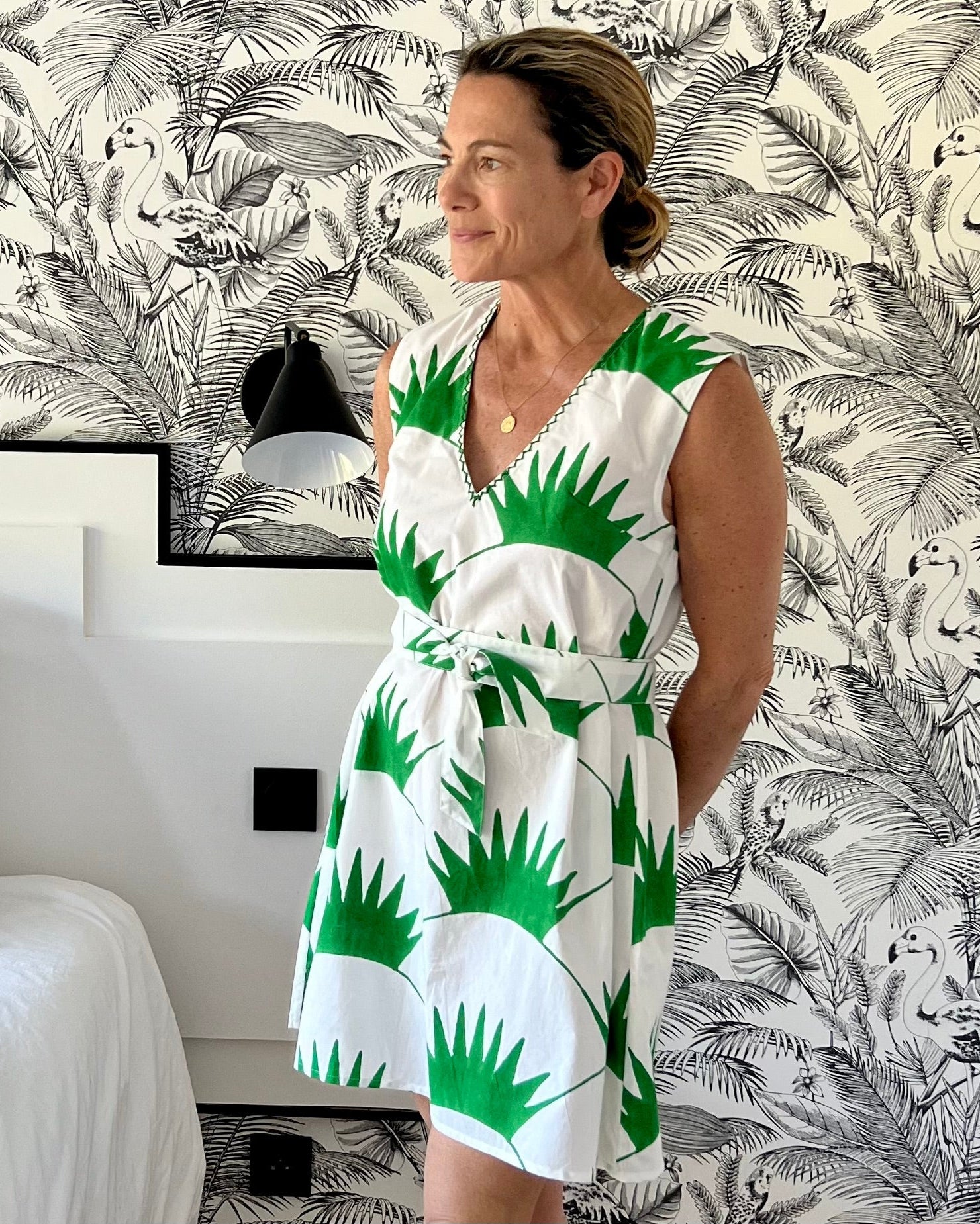 A woman standing in a room with a sleeveless shift dress featuring a grass green print on a white background