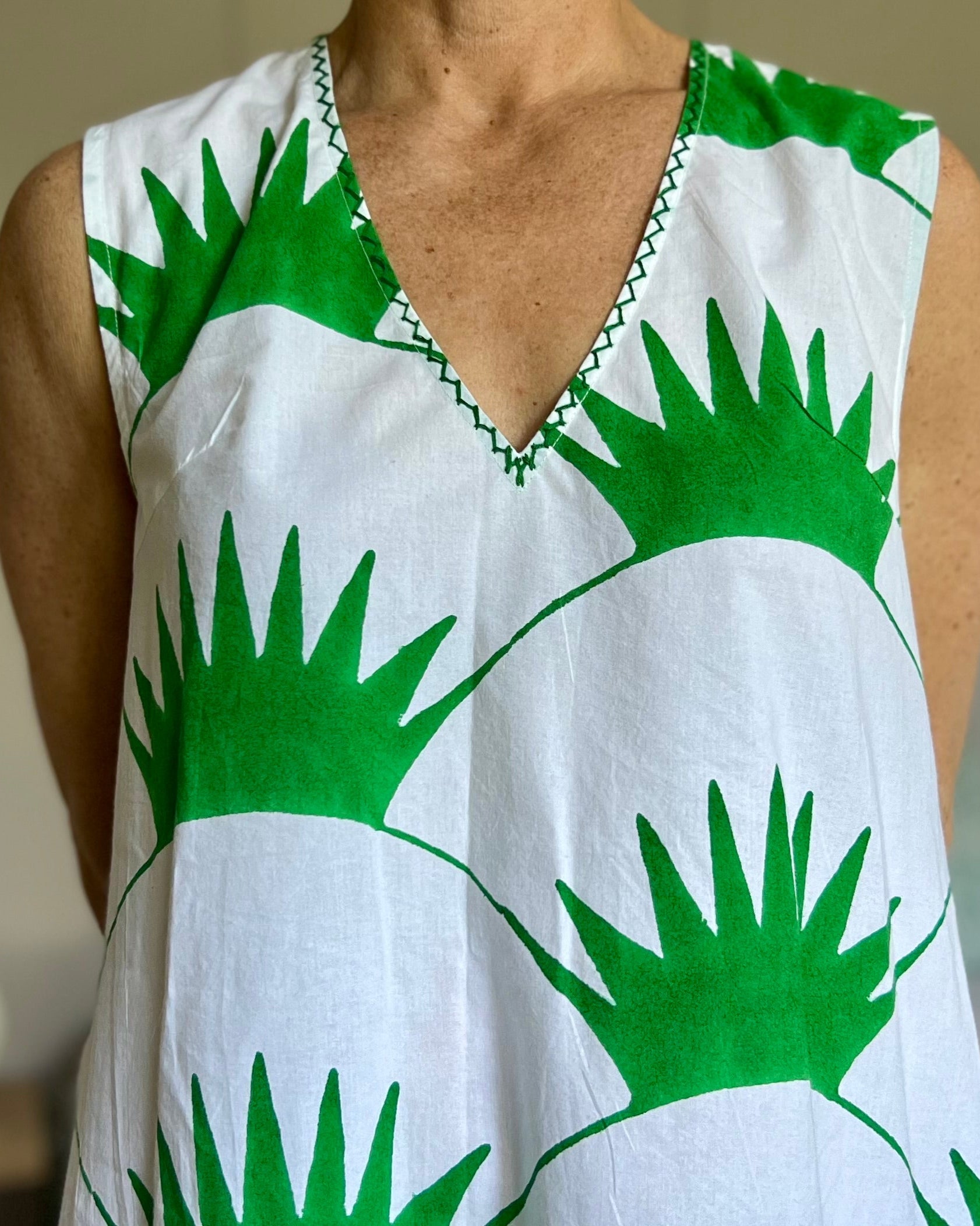 A woman standing in a room with a sleeveless shift dress featuring a grass green print on a white background