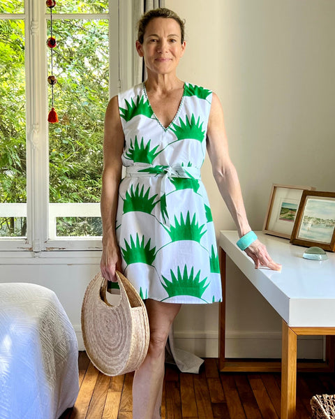 A woman standing in a room with a sleeveless shift dress featuring a grass green print on a white background, paired with a straw bag and white sandals.