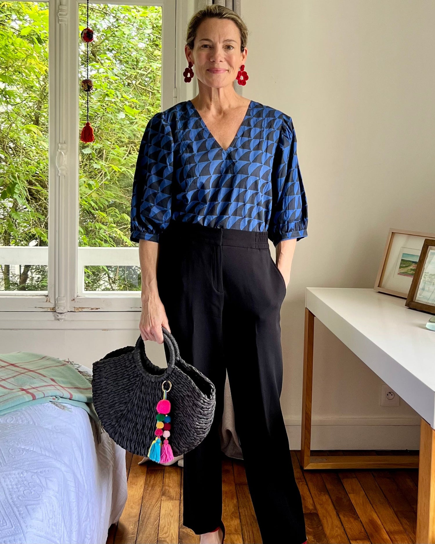 A person wearing a navy blue and black blouse with a deep-V neck and 3/4 length puff sleeves, paired with black trousers, standing in a room with a bed and a desk in the background.
