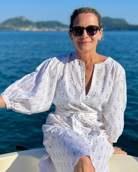 A woman sitting on a boat wearing a white puff sleeve dress with blue print posing with the ocean and hills in the background.