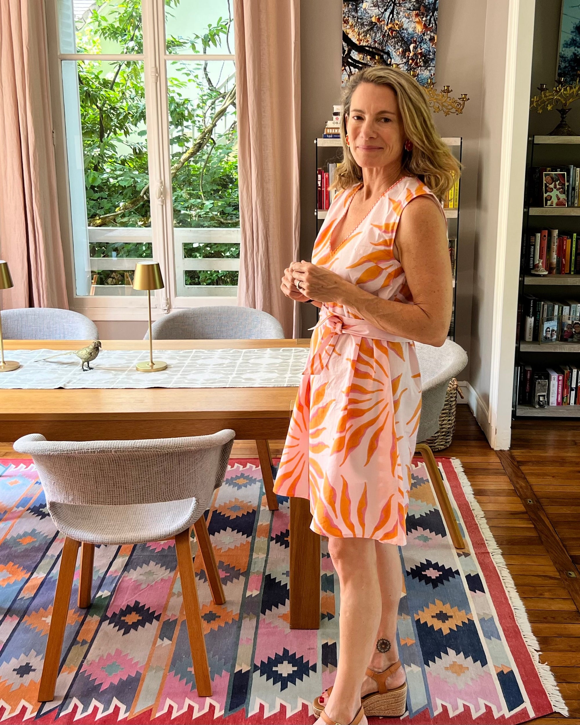Woman in a pink and orange hand block-printed organic cotton dress standing in a dining room with a colorful rug and bookshelf.