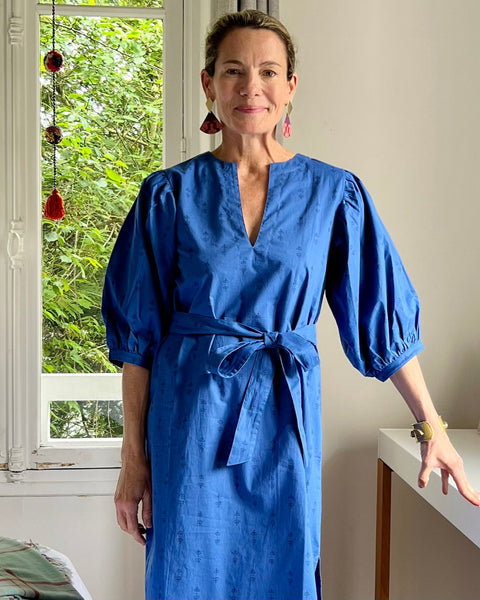 A woman standing in a room wearing a royal blue dress with puff sleeves, a V-neck, and a sash belt.