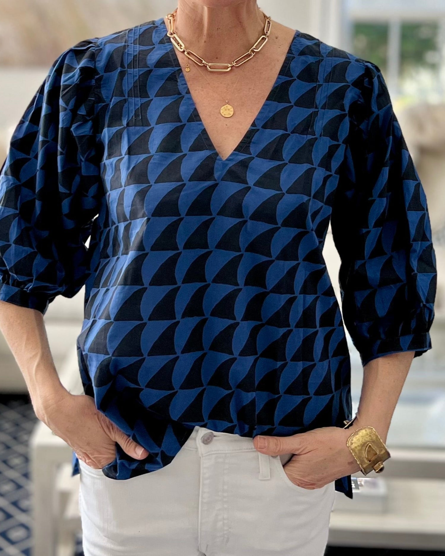 A person wearing a navy blue and black blouse with a deep-V neck and 3/4 length puff sleeves in white jeans in a light-filled sitting room.