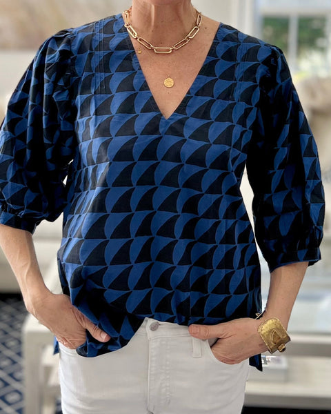 A person wearing a navy blue and black blouse with a deep-V neck and 3/4 length puff sleeves in white jeans in a light-filled sitting room.